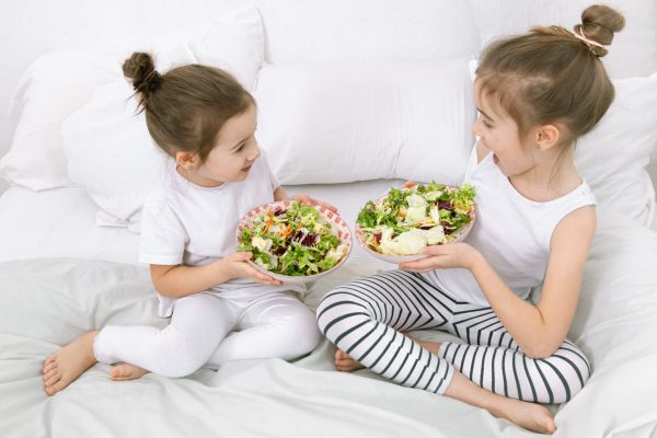 Healthy food at home. Happy two cute children eating fruits and vegetables in the bedroom on the bed. Healthy food for children and teenagers.